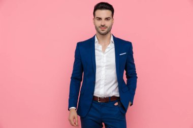 Pozitif iş adamı elleri cebinde gülümserken mavi takım elbise giyer ve pembe stüdyo arka planında durur.