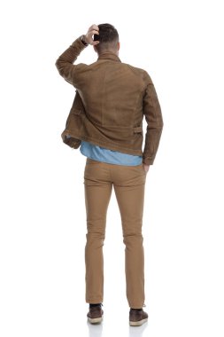 Deri ceket giyip beyaz stüdyo arka planında dikilen düşünceli, sıradan bir adamın kafasını kaşıması.