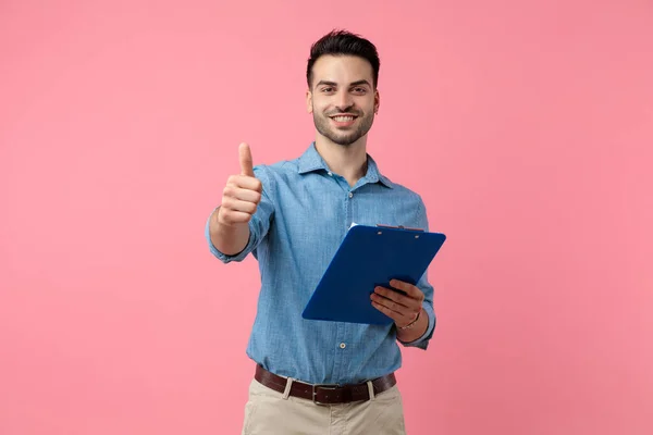 快乐的年轻人拿着剪贴板 竖起大拇指 站在粉色的背景上 — 图库照片
