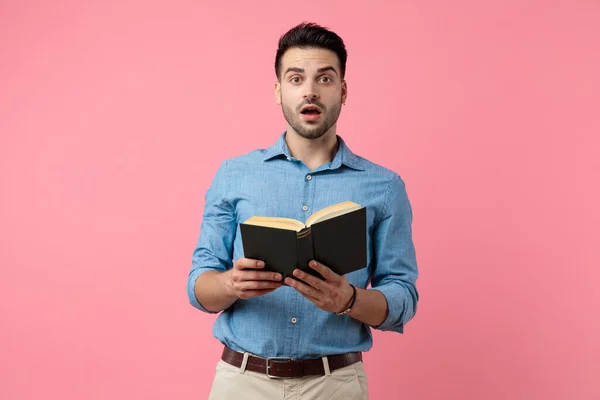 ショックを受けた若いカジュアルな男が本を読んでピンクの背景に立って — ストック写真