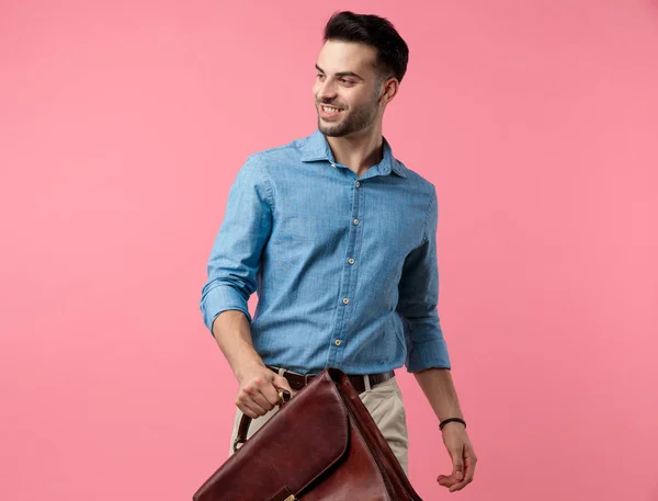 happy young guy in denim shirt smiling and holding suitcase, standing on pink background