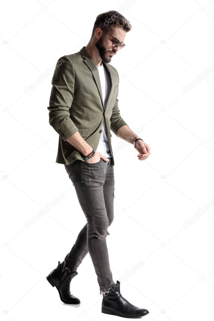 Side view of a thoughtful model holding his hand in his pocket while wearing green jacket and sunglasses, walking on white studio background