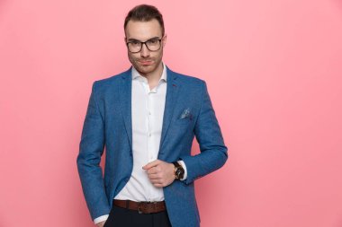 Pembe stüdyo arka planında dururken ceketini ayarlayan ve gülümseyen zeki bir adam.