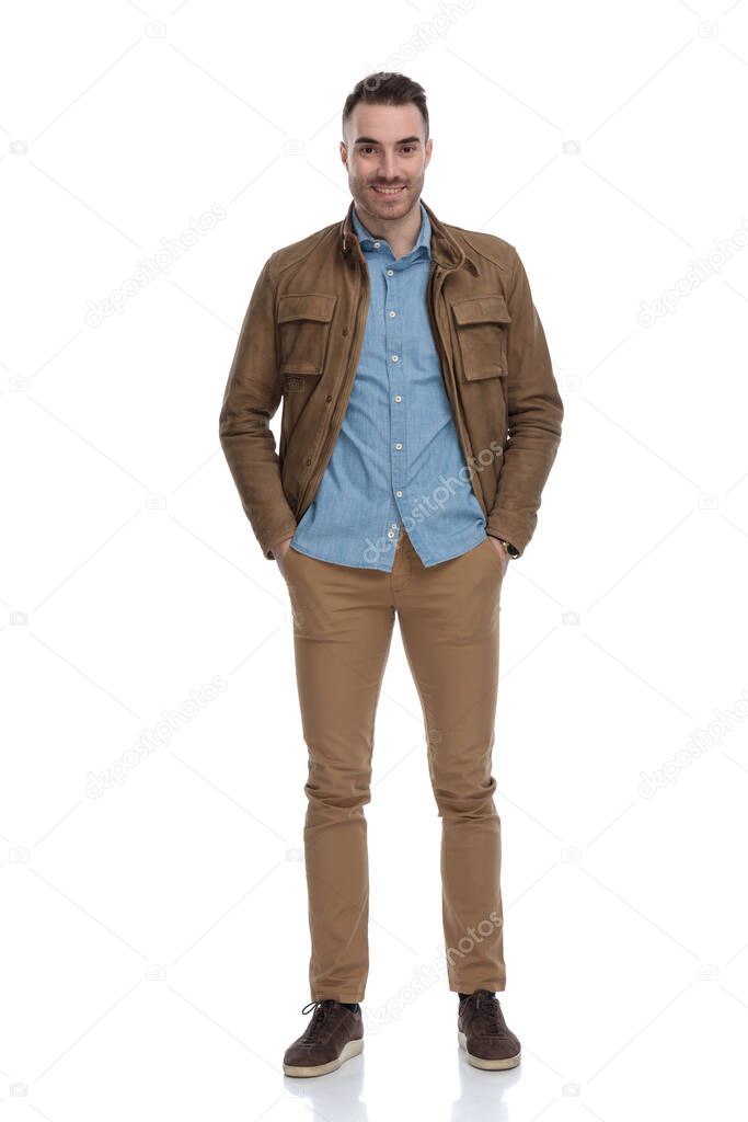 Happy casual man holding both hands in his pockets, smiling while wearing a leather jacket and stepping on white studio background