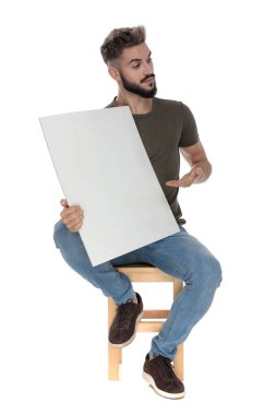 Dost canlısı sıradan bir adam boş bir ilan panosunu sunuyor, beyaz stüdyo arka planında bir sandalyede oturuyor.