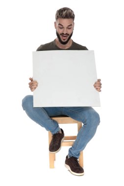 Şaşırmış sıradan bir adam soluk soluğa kalmış ve boş bir reklam panosuna bakıyor, beyaz stüdyo arka planında bir sandalyede oturuyor.