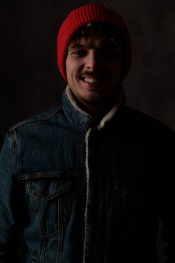 Kırmızı şapkalı ve kot ceketli mutlu adam, gülüyor ve siyah arka planda poz veriyor.