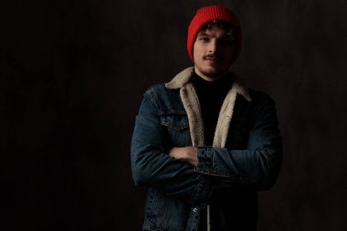 Mutlu, havalı bir model kot ceket ve kırmızı şapka giyiyor, kollarını kavuşturup gülümsüyor, siyah arka planda duruyor.