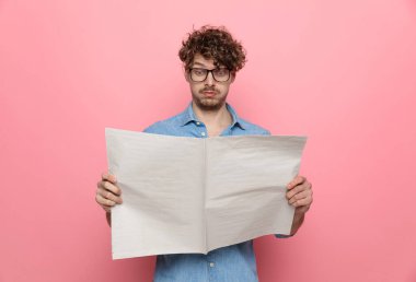 Kot gömlekli genç adam gazete okuyor, pembe arka planda duruyor.