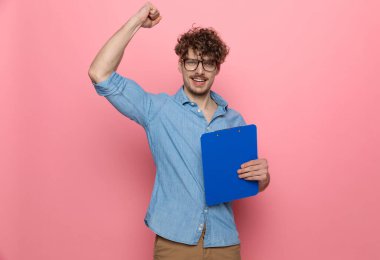 Genç, sıradan bir adam elinde bir pano, zaferi kutluyor, yumruğunu havada tutuyor, pembe arka planda duruyor.