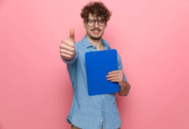 Genç, sıradan bir adam elinde bir not panosu tutuyor ve pembe arka planda başparmak işareti yapıyor.