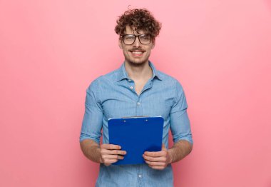 Elinde pano olan gülümseyen genç adam sunuyor, pembe arka planda duruyor.