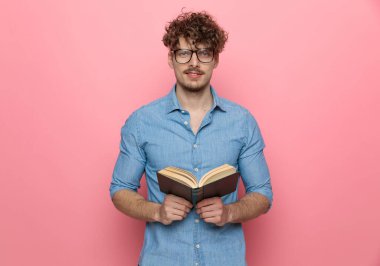 Sırıtan, elinde kitap tutan, pembe arka planda duran genç, seksi, sıradan bir adam.