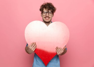 Mutlu genç adam büyük kırmızı bir kalp tutuyor ve gülümsüyor, pembe arka planda duruyor.