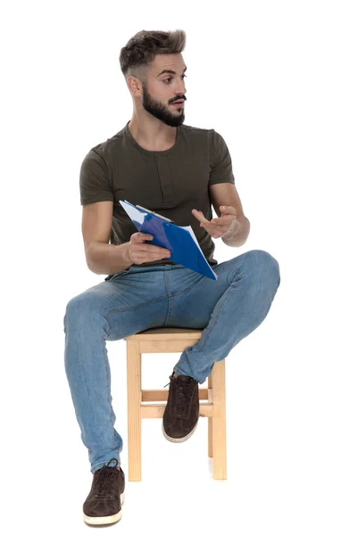 一个英俊的学生坐在白色的工作室背景的椅子上 一边指指点点一边谈论他的记事板和笔记 — 图库照片