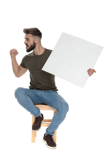 白いスタジオの背景にある椅子に座って 白い看板を保持しながら怒っているカジュアルな男は叫んでジェスチャー — ストック写真
