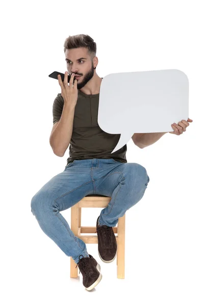 白いスタジオの背景にある椅子に座って電話で話して 空白のスピーチバブルを保持しているハンサムなカジュアルな男 — ストック写真