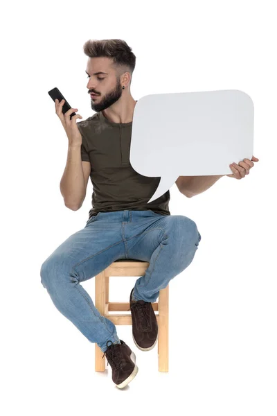 Przystojny Facet Patrząc Telefon Trzymając Puste Mowy Bańki Siedzi Krześle — Zdjęcie stockowe