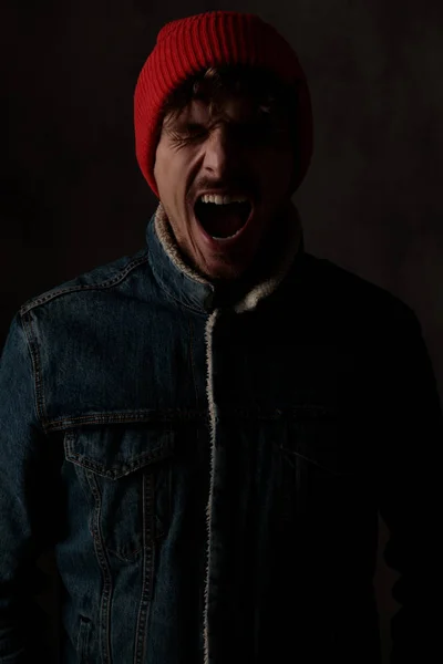 赤い帽子とデニムのジャケットを身に着けて黒の背景で叫んで目を閉じる劇的なクールな男 — ストック写真