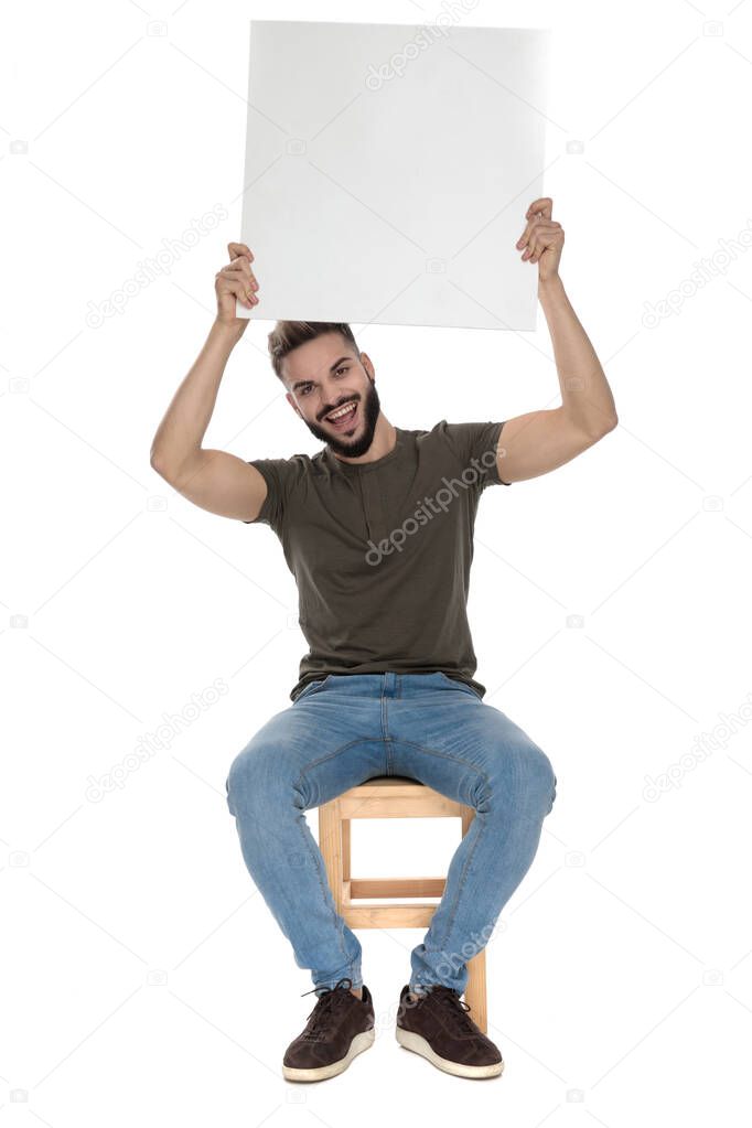Cheerful casual man holding a blank billboard and laughing, sitting on a chair on white studio background