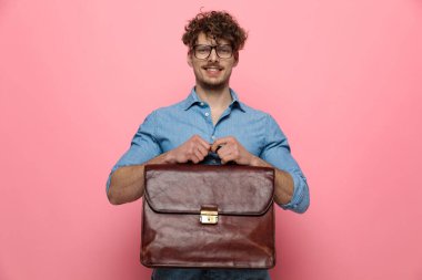 Mutlu genç adam gülümsüyor ve bavulu tutuyor, pembe arka planda duruyor.