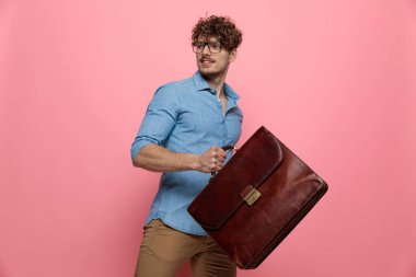 Yanlara bakan, bavulu tutan, pembe arka planda yürüyen seksi genç adam.