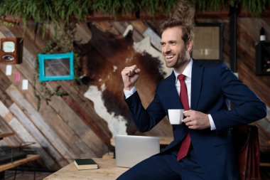 Heyecanlı iş adamı kahve içiyor, gülüyor, yumruğunu havaya kaldırıyor ve zaferi kahvecide kutluyor.