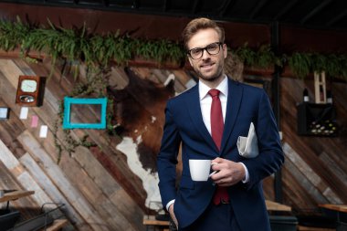 Genç işadamı elinde gazete tutuyor, kahve içiyor ve gülümsüyor, kahve dükkanında duruyor.