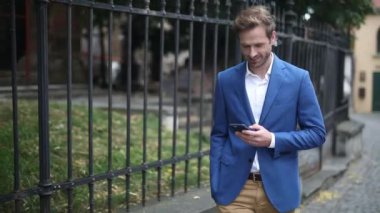 Çekici iş adamı donanma takımı giyiyor telefonda mutlu bir şekilde mesajlaşırken el ele yürüyor ve sonra dışarıda tellerin yanında telefonla konuşuyor.