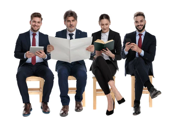 该小组由4名商人组成 他们在等待求职面试时 从书本 电话和平板电脑中阅读 他们坐在有白色工作室背景的椅子上 — 图库照片
