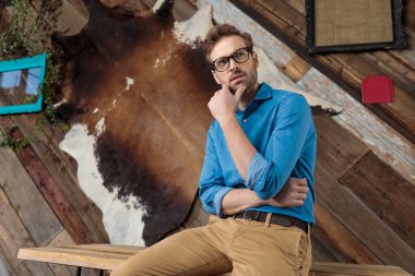 Mavi gömlek ve gözlük giyerken, kahve fonu üzerinde bir masanın üzerinde otururken, düşünceli manken bakışlarını kaçırıp çenesine dokunuyor.