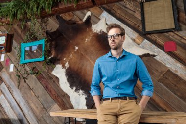 Rahatsız bir model gözlerini kaçırıyor ve mavi gömlek ve gözlük takıp kahve fonu üzerinde bir masada otururken iki elini cebine sokuyor.