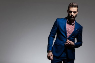 Yakışıklı iş adamı donanma takımı giyiyor ve gri stüdyo arka planında ciddi bir şekilde ceketin düğmesinin üzerinde duruyor ve el ele tutuşuyor.