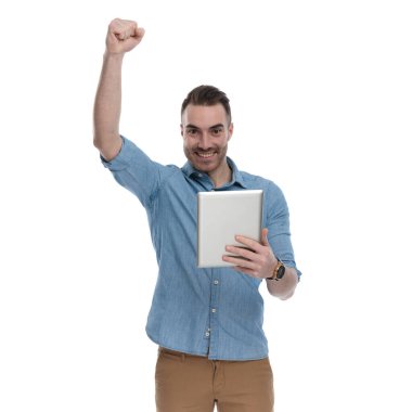 Tablet tutan neşeli, sıradan bir adam mavi gömlek giyerken kutlama yapıyor, beyaz stüdyo arka planında duruyor.