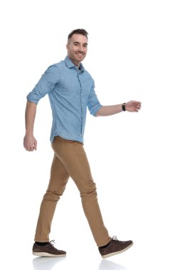 Gülümseyen, mavi gömlekli, beyaz stüdyo arka planında yürüyen mutlu sıradan bir adamın yan görüntüsü.