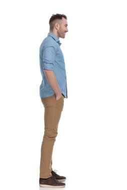 Mavi gömlekli, beyaz stüdyo arka planında duran ve elleri ceplerinde gülümseyen sıradan bir adamın yan görüntüsü.