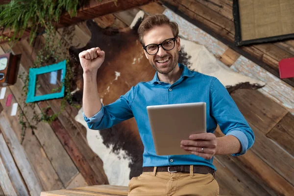 快乐模特拿着平板电脑 戴着蓝色衬衫和眼镜 坐在咖啡后的桌子上庆祝胜利 — 图库照片