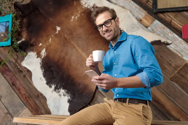 Positive model smiling, holding coffee cup and phone while wearing blue shirt and glasses, sitting on a desk on coffeeshop background