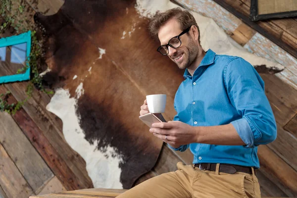 青いシャツと眼鏡をかけながら コーヒーカップを持ち 電話を見て コーヒーショップの背景にある机の上に座って笑っている幸せなモデル — ストック写真