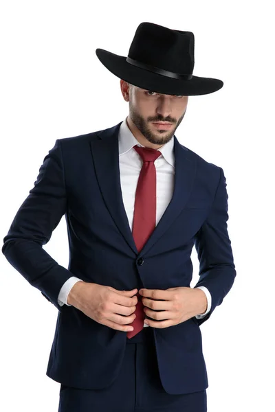 帽子とスーツを身に着けている間に彼のスーツを修正しながら 真剣にビジネスマンを見て 白いスタジオの背景に立って — ストック写真