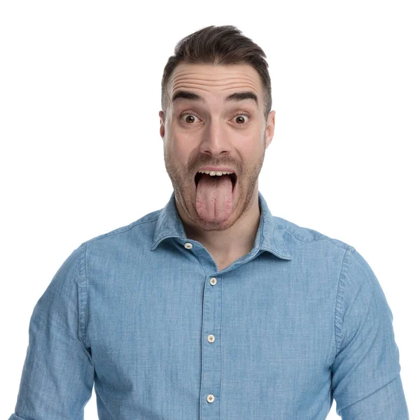 穿着蓝色衬衫 站在白工作室背景上 笨手笨脚 随和的男人伸出舌头 — 图库照片