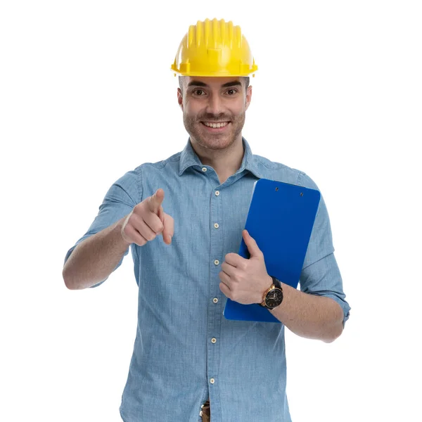Szczęśliwy Człowiek Casual Wskazując Trzymając Schowek Nosząc Kask Bezpieczeństwa Stojąc — Zdjęcie stockowe