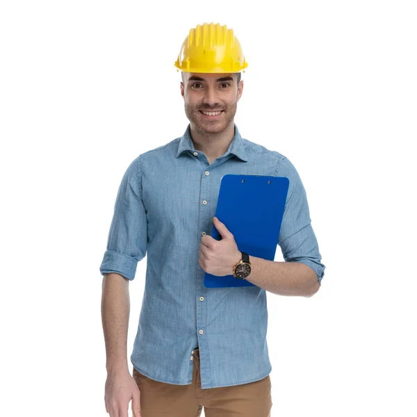快乐而随意的男人拿着剪贴板 头戴安全帽 站在白色的工作室背景上 — 图库照片