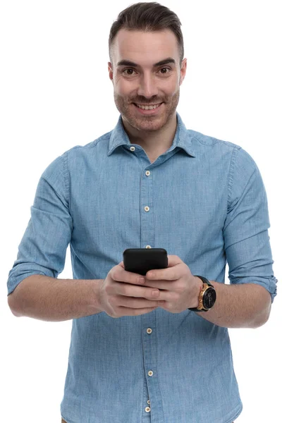快乐而随意的男人拿着电话 穿着蓝色衬衫 站在白色的工作室背景上 微笑着 — 图库照片