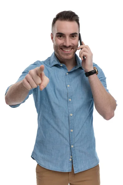 快乐而随意的男人 穿着蓝色衬衫 站在白色的工作室背景上 一边打电话一边笑一边指指点点 — 图库照片