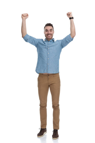 Cheerful casual man celebrating with both fists in the air while wearing blue shirt, standing on white studio background