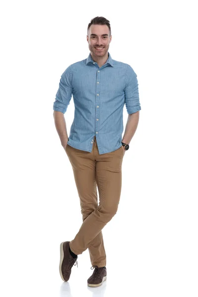 白いスタジオの背景に立って 青いシャツを着ている間 彼のポケットに両手で笑みを浮かべて幸せなカジュアル男 — ストック写真