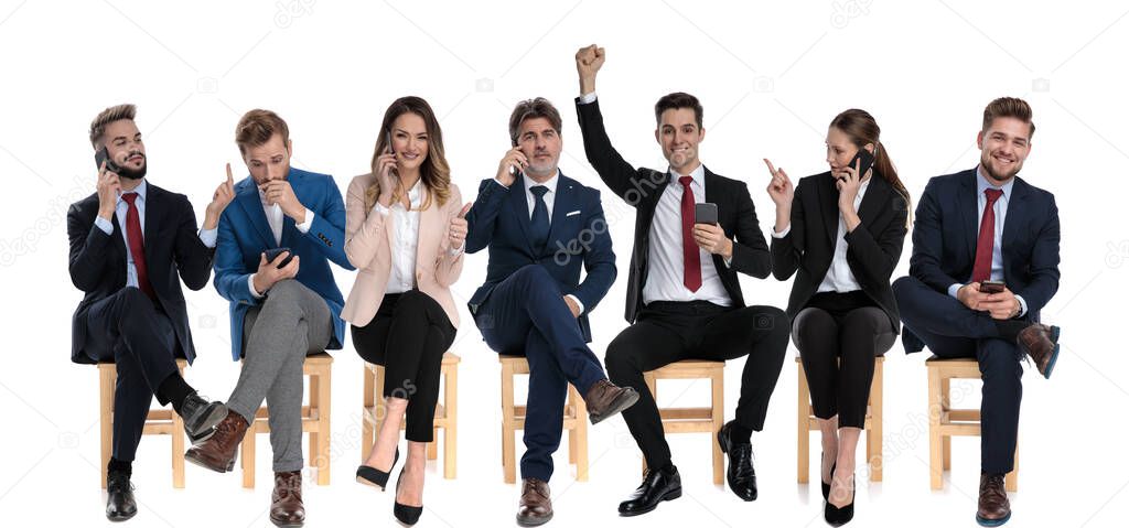 Team of 7 businessmen celebrating and talking on their phones while one is giving the middle finger,  sitting on chairs on white studio background