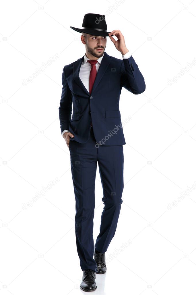 Eager businessman looking away and fixing his hat while wearing suit, stepping on white studio background