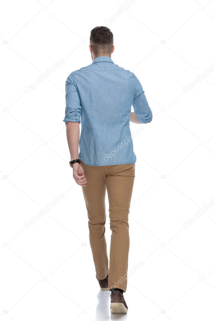Rear view of confident casual man wearing blue shirt, walking on white studio background